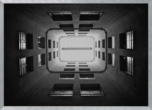 A symmetrical, black and white photograph looking directly up from the base of a building's interior courtyard. The dark walls rise up, lined with rows of windows, creating a powerful sense of perspective that draws the eye to the bright rectangle of the sky or a skylight at the top. The image is enclosed in a simple silver frame. Poster