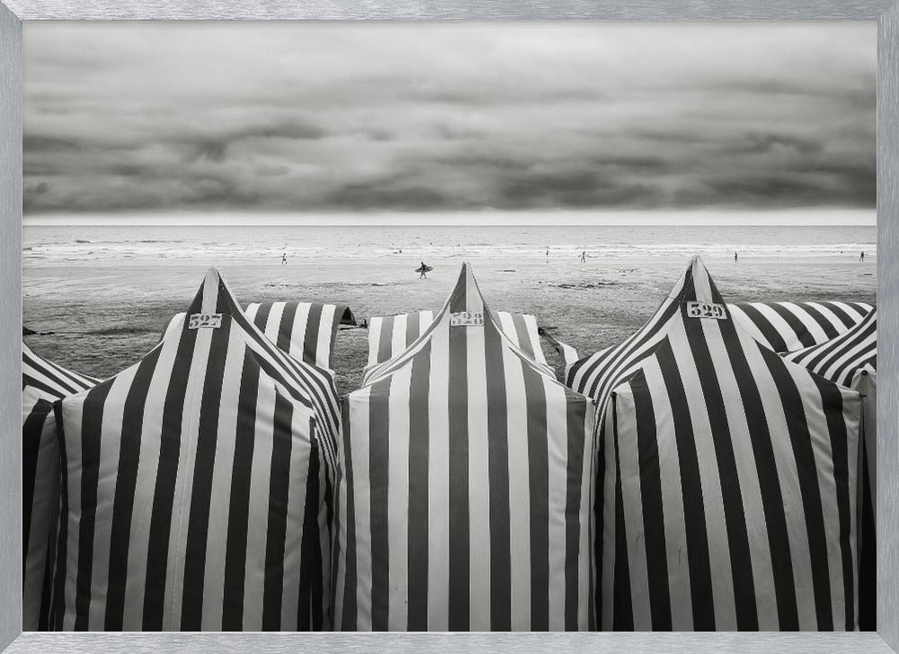 A framed, black and white photograph of striped beach tents on a cloudy day, with the ocean and distant surfers in the background. Print