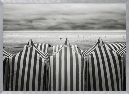A framed, black and white photograph of striped beach tents on a cloudy day, with the ocean and distant surfers in the background. Print