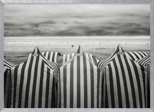 A framed, black and white photograph of striped beach tents on a cloudy day, with the ocean and distant surfers in the background. Print