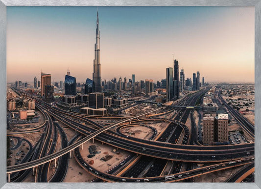 An aerial photograph of the Dubai skyline at sunset, featuring the iconic Burj Khalifa towering over other skyscrapers, with a complex web of intersecting highways and overpasses in the foreground, all under a gradient sky of orange and blue. The photo is displayed within a silver frame. Poster