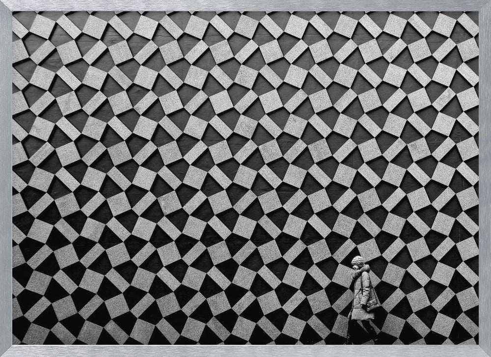 A black and white photograph of a lone person walking in front of a massive wall covered in a dizzying geometric pattern of tilted, textured gray quadrilaterals on a black background. The figure in the bottom right corner is dwarfed by the scale of the abstract architectural design. The image has a silver-gray frame. Artwork