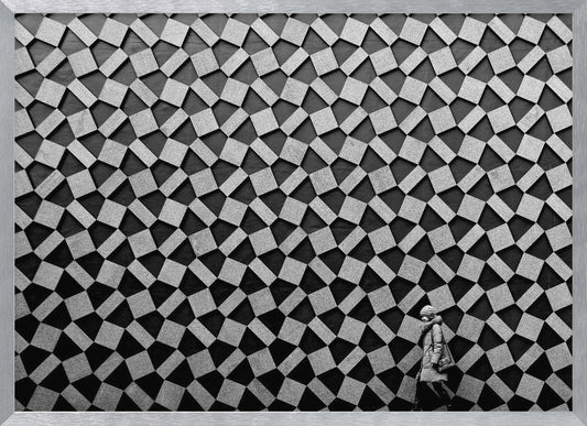 A black and white photograph of a lone person walking in front of a massive wall covered in a dizzying geometric pattern of tilted, textured gray quadrilaterals on a black background. The figure in the bottom right corner is dwarfed by the scale of the abstract architectural design. The image has a silver-gray frame. Artwork