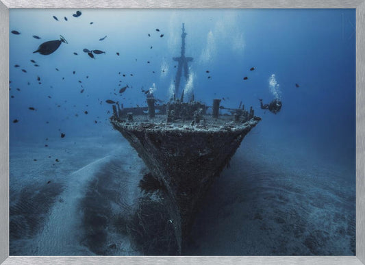 An underwater photograph of a large, sunken shipwreck resting on the sandy ocean floor. Two scuba divers are exploring the wreck, surrounded by schools of small fish in the deep blue water, with light filtering down from the surface. Print