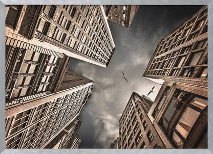 A dramatic, low-angle photograph looking up at the sky between several tall, sepia-toned skyscrapers that converge towards the center. Two birds are seen flying in the moody, dark grey sky. The image has a vintage feel and is presented in a silver frame. Wall Art