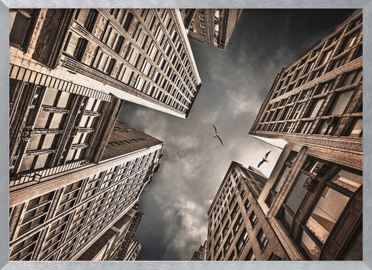 A dramatic, low-angle photograph looking up at the sky between several tall, sepia-toned skyscrapers that converge towards the center. Two birds are seen flying in the moody, dark grey sky. The image has a vintage feel and is presented in a silver frame. Wall Art