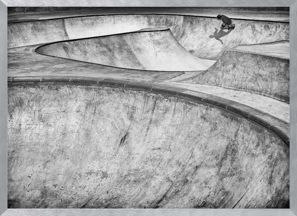 A high-angle, black and white photograph of a sprawling concrete skatepark with smooth, curving ramps. A single skateboarder is visible in the upper right, casting a distinct shadow on the ramp as they ride. The image is enclosed in a silver frame. Decor