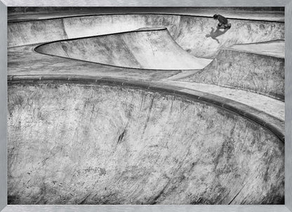 A high-angle, black and white photograph of a sprawling concrete skatepark with smooth, curving ramps. A single skateboarder is visible in the upper right, casting a distinct shadow on the ramp as they ride. The image is enclosed in a silver frame. Decor