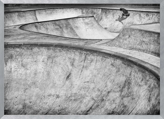 A high-angle, black and white photograph of a sprawling concrete skatepark with smooth, curving ramps. A single skateboarder is visible in the upper right, casting a distinct shadow on the ramp as they ride. The image is enclosed in a silver frame. Decor