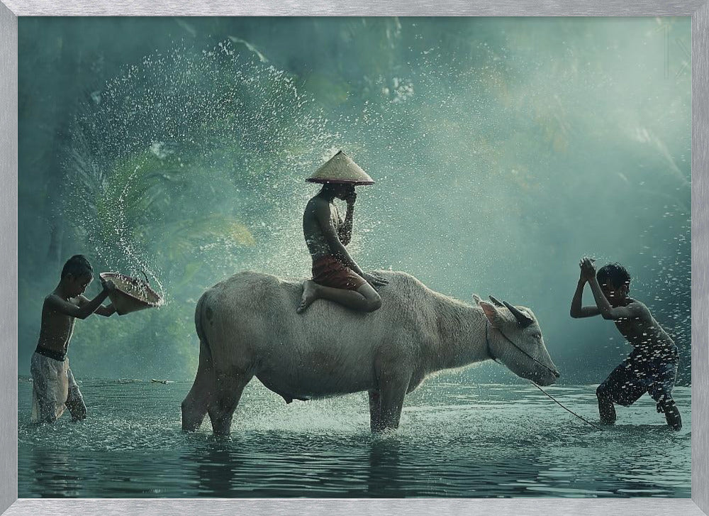 A serene photograph of three young boys playing in a river with a large water buffalo, one boy sits atop the animal while the others splash water, all set against a misty green backdrop and framed in silver. Print