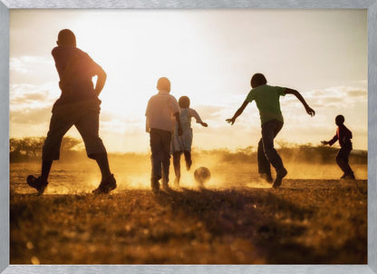 A low-angle, warmly lit photograph of five children silhouetted against a bright sunset as they play soccer on a dusty field, kicking up clouds of dust in the golden light. Decor