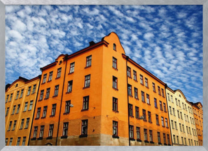 A low-angle shot of a vibrant orange apartment building against a dramatic blue sky filled with small, puffy white clouds. The building is on a corner, with another yellow-hued building visible next to it. The entire image is enclosed in a silver, brushed-metal style frame. Artwork