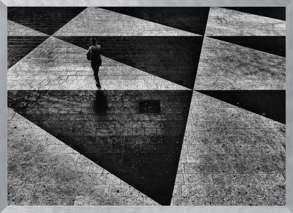A dramatic, high-contrast black and white overhead photograph of a person walking across a paved ground with a striking geometric pattern of light and dark triangles created by strong shadows. The lone figure casts a long shadow, enhancing the minimalist and abstract composition, which is enclosed in a grey frame. Print