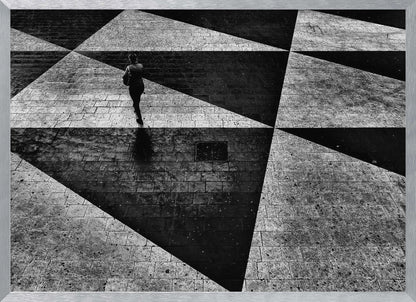 A dramatic, high-contrast black and white overhead photograph of a person walking across a paved ground with a striking geometric pattern of light and dark triangles created by strong shadows. The lone figure casts a long shadow, enhancing the minimalist and abstract composition, which is enclosed in a grey frame. Print
