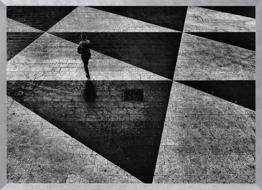 A dramatic, high-contrast black and white overhead photograph of a person walking across a paved ground with a striking geometric pattern of light and dark triangles created by strong shadows. The lone figure casts a long shadow, enhancing the minimalist and abstract composition, which is enclosed in a grey frame. Print