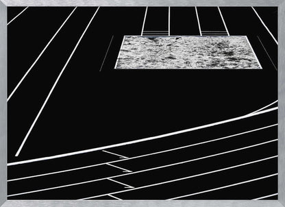 A high-contrast, black and white abstract artwork depicting a running track from an elevated perspective. Stark white lines representing lanes curve across a solid black background. In the upper portion, a rectangular shape filled with a textured, water-like pattern replaces a typical sand pit. The entire piece is presented within a silver frame. Print