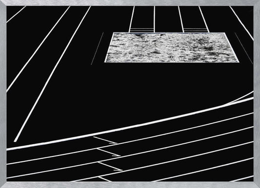 A high-contrast, black and white abstract artwork depicting a running track from an elevated perspective. Stark white lines representing lanes curve across a solid black background. In the upper portion, a rectangular shape filled with a textured, water-like pattern replaces a typical sand pit. The entire piece is presented within a silver frame. Print