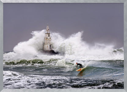 A framed photograph of a surfer in a black wetsuit riding a wave in a stormy sea. In the background, massive white waves crash against an old white lighthouse under a grey, overcast sky. Artwork