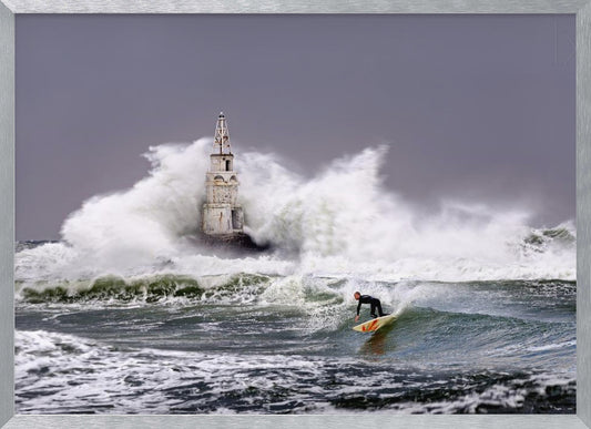 A framed photograph of a surfer in a black wetsuit riding a wave in a stormy sea. In the background, massive white waves crash against an old white lighthouse under a grey, overcast sky. Artwork
