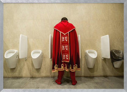 A back view of a person wearing an elaborate, long red and gold cape, standing at a urinal in a modern public restroom with beige tiled walls and multiple white urinals. Artwork