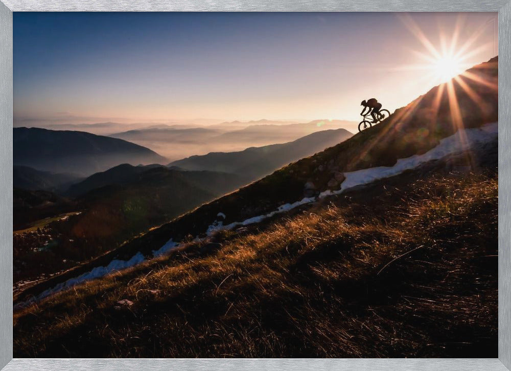A silhouette of a mountain biker pushing their bike up a steep mountainside against a brilliant sunrise. The sun is cresting the peak, creating a starburst effect with long light rays. In the background, layers of mountains and valleys are filled with a soft morning mist under a clear gradient sky. Artwork