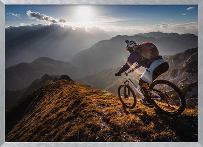 A mountain biker navigates a narrow, golden-lit ridge against a backdrop of a vast mountain range. The sun is breaking through clouds in the distance, casting dramatic rays of light across the epic landscape. Print