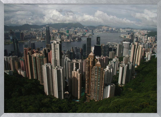 A high-angle photograph of the Hong Kong cityscape, viewed from a lush green peak. Numerous skyscrapers fill the frame, overlooking a bustling harbor with mountains in the distant background under a cloudy sky. The entire image is presented within a silver, brushed-metal frame. Artwork