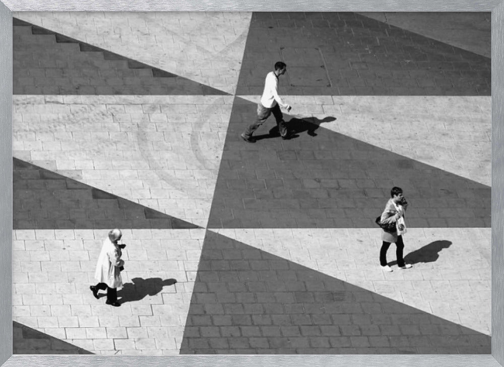 A high-angle, black and white photograph capturing three people walking across a paved plaza, their paths intersecting with a dramatic geometric pattern of light and shadow. The strong sunlight creates large, sharp-edged triangles of shade, isolating each figure in the composition, which is enclosed in a silver frame. Decor