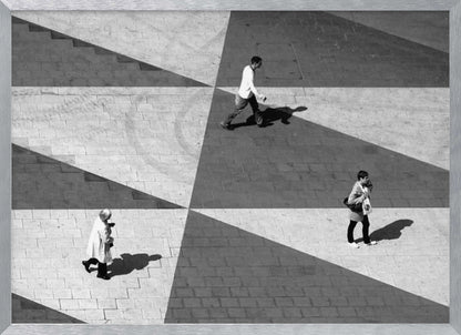 A high-angle, black and white photograph capturing three people walking across a paved plaza, their paths intersecting with a dramatic geometric pattern of light and shadow. The strong sunlight creates large, sharp-edged triangles of shade, isolating each figure in the composition, which is enclosed in a silver frame. Decor