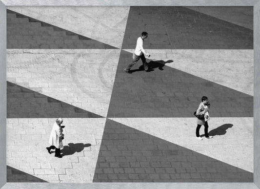 A high-angle, black and white photograph capturing three people walking across a paved plaza, their paths intersecting with a dramatic geometric pattern of light and shadow. The strong sunlight creates large, sharp-edged triangles of shade, isolating each figure in the composition, which is enclosed in a silver frame. Decor