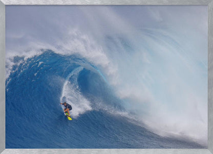 A framed photograph of a professional surfer skillfully riding down the steep face of a massive, cresting blue ocean wave, surrounded by white sea spray. Decor