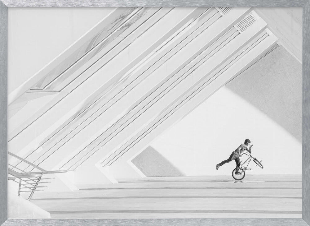 A minimalist black and white photograph in a silver frame, featuring a lone BMX rider performing a trick on the back wheel inside a stark, modern building with strong diagonal architectural lines. Poster