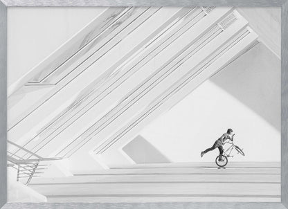 A minimalist black and white photograph in a silver frame, featuring a lone BMX rider performing a trick on the back wheel inside a stark, modern building with strong diagonal architectural lines. Poster
