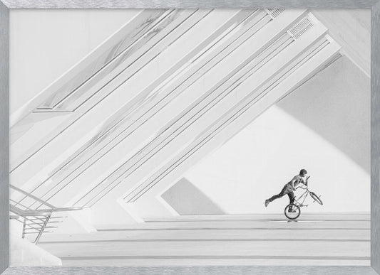 A minimalist black and white photograph in a silver frame, featuring a lone BMX rider performing a trick on the back wheel inside a stark, modern building with strong diagonal architectural lines. Poster