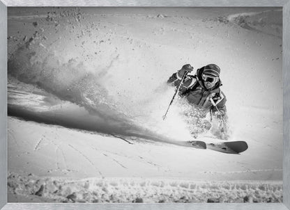 A framed, black and white action shot of a skier making a sharp turn on a snowy slope, sending a large spray of powder into the air. The skier is leaning into the turn, fully kitted out in winter gear including goggles and a beanie. Artwork