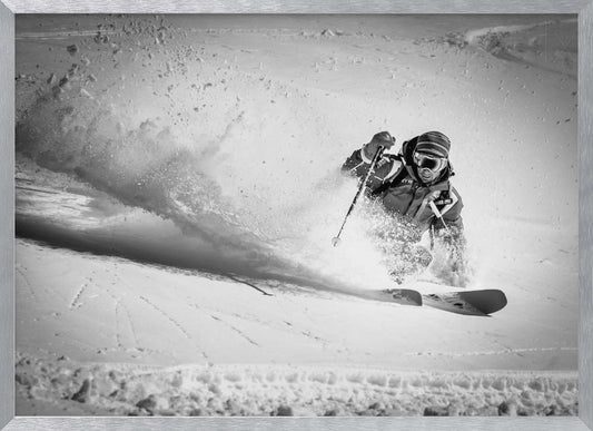 A framed, black and white action shot of a skier making a sharp turn on a snowy slope, sending a large spray of powder into the air. The skier is leaning into the turn, fully kitted out in winter gear including goggles and a beanie. Artwork