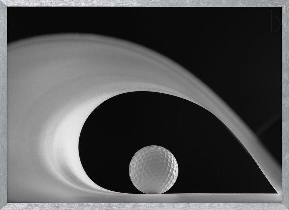 A dramatic black and white still life photo featuring a golf ball set against a stark black background. A large, abstract white shape swirls gracefully over the ball, creating a dynamic, wave-like composition. The image is presented within a silver frame. Decor