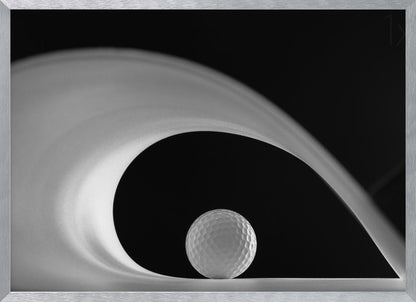 A dramatic black and white still life photo featuring a golf ball set against a stark black background. A large, abstract white shape swirls gracefully over the ball, creating a dynamic, wave-like composition. The image is presented within a silver frame. Decor
