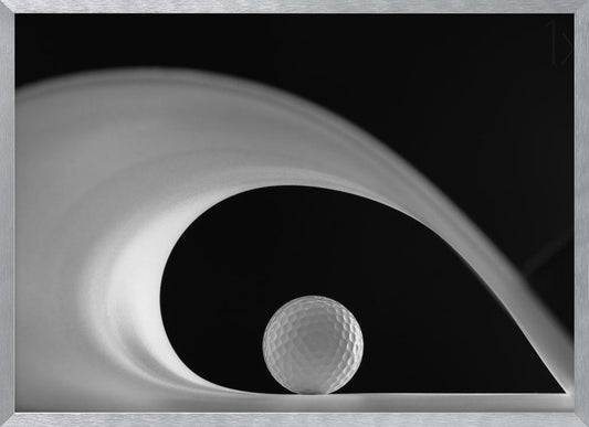 A dramatic black and white still life photo featuring a golf ball set against a stark black background. A large, abstract white shape swirls gracefully over the ball, creating a dynamic, wave-like composition. The image is presented within a silver frame. Decor
