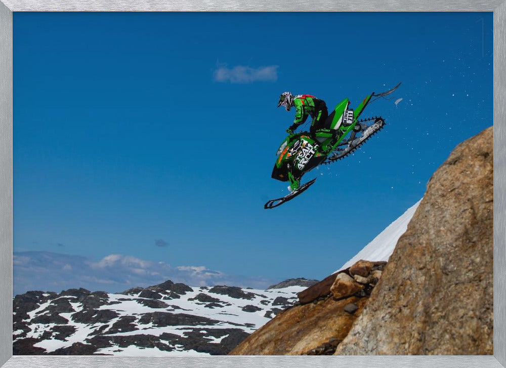 A dynamic action shot of a person in full gear launching a green snowmobile into the air off a rocky, snow-covered cliff, set against a backdrop of a vivid blue sky and distant snowy mountains. The image is enclosed in a silver frame. Wall Art
