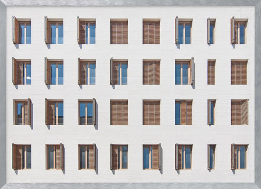 A symmetrical architectural photo of a modern white building facade featuring a grid of twenty-eight windows with wooden louvered shutters, some open and some closed, creating a varied pattern, all within a brushed silver frame. Decor