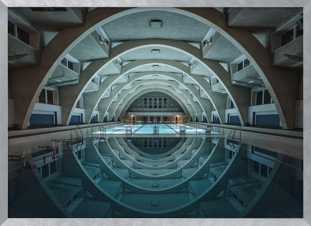 An atmospheric photograph of an empty indoor swimming pool with remarkable Brutalist-style architecture. A series of large, repeating concrete arches span the length of the pool, creating a tunnel-like effect. The arches and lights are perfectly reflected in the still, blue water, forming a symmetrical, eye-like shape. Decor