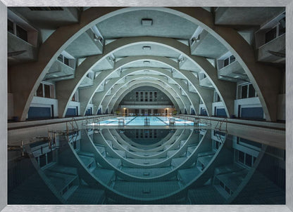 An atmospheric photograph of an empty indoor swimming pool with remarkable Brutalist-style architecture. A series of large, repeating concrete arches span the length of the pool, creating a tunnel-like effect. The arches and lights are perfectly reflected in the still, blue water, forming a symmetrical, eye-like shape. Decor