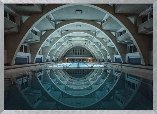 An atmospheric photograph of an empty indoor swimming pool with remarkable Brutalist-style architecture. A series of large, repeating concrete arches span the length of the pool, creating a tunnel-like effect. The arches and lights are perfectly reflected in the still, blue water, forming a symmetrical, eye-like shape. Decor