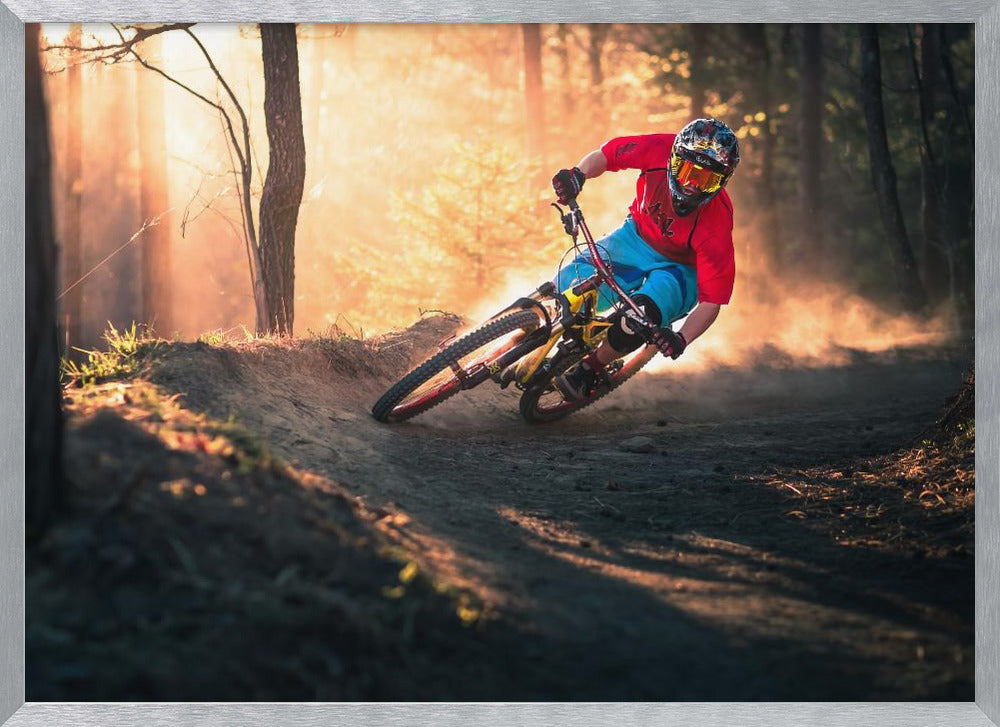 A mountain biker in a red shirt, blue shorts, and full-face helmet leans sharply into a dusty turn on a dirt trail in a forest. The setting sun casts a warm, golden light through the trees, creating a dramatic, backlit effect. Wall Art