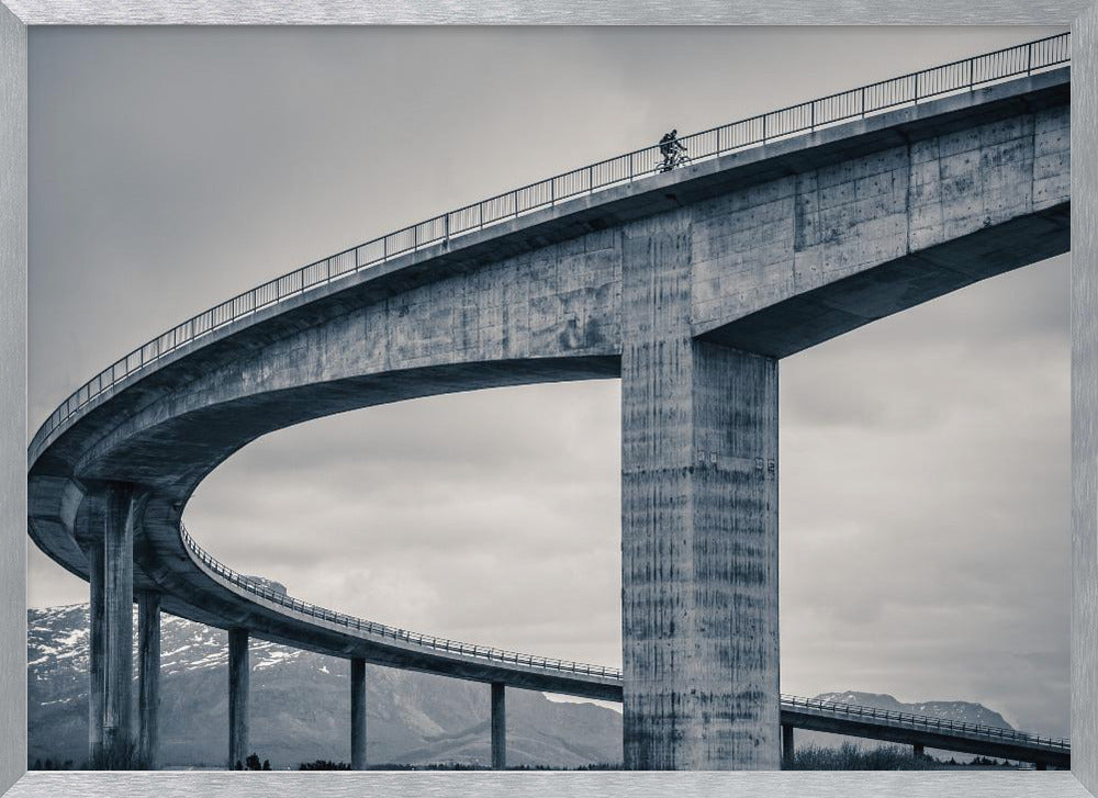 A black and white photograph of a lone cyclist riding across the top level of a massive, curving concrete bridge, which sweeps across the frame. In the background, a lower level of the bridge and distant mountains are visible under a cloudy sky. Decor