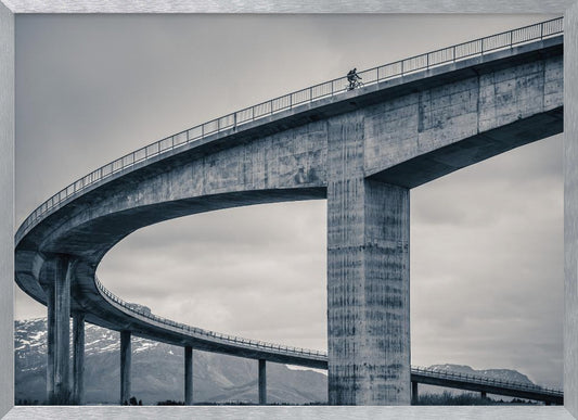 A black and white photograph of a lone cyclist riding across the top level of a massive, curving concrete bridge, which sweeps across the frame. In the background, a lower level of the bridge and distant mountains are visible under a cloudy sky. Decor