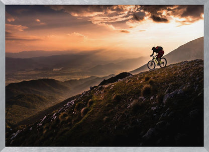 A lone mountain biker is silhouetted against a breathtaking sunset as they navigate a steep, rocky ridge. Below them, layers of mountains and a vast valley stretch into the distance under a dramatic, cloudy orange sky. Artwork
