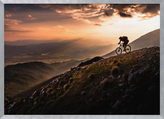 A lone mountain biker is silhouetted against a breathtaking sunset as they navigate a steep, rocky ridge. Below them, layers of mountains and a vast valley stretch into the distance under a dramatic, cloudy orange sky. Artwork
