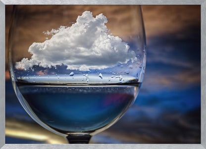 A surreal close-up of a wine glass holding blue liquid and a single white cloud, with water droplets on the glass, set against a blurred background and enclosed in a silver frame. Print
